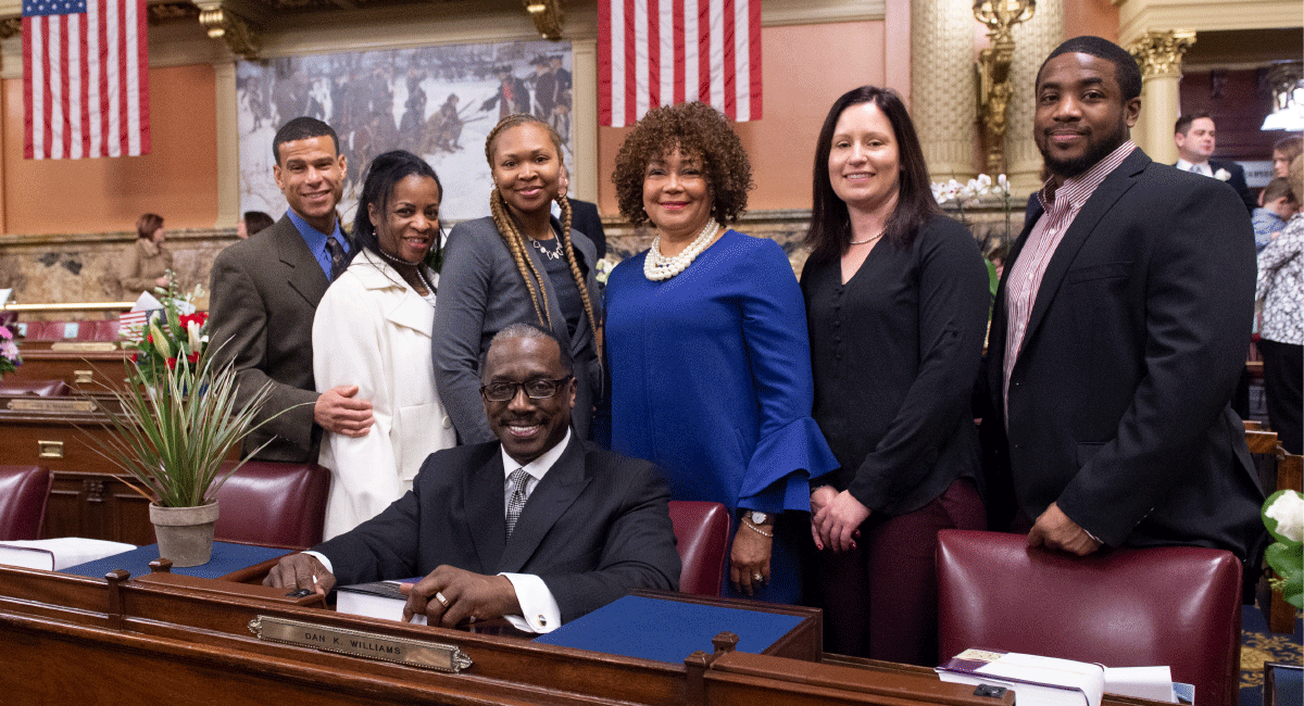 Representative Williams sitting and posing for photo with group at his seat on House floor